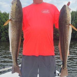 Man in bright shirt holds two large fish on a boat with water and trees in the background.