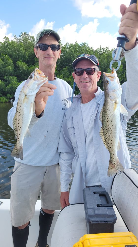 Two men smiling on a boat, holding caught fish in each hand against a backdrop of greenery.