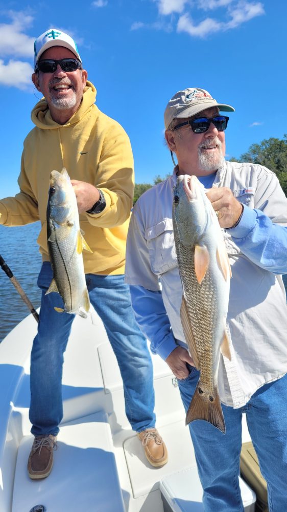 Two men on a boat holding fish, smiling under a clear blue sky.