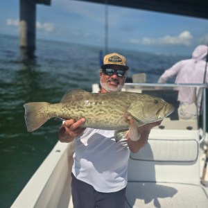Man in hat holds large fish on a boat under a bridge, with water and another person in the background.