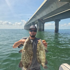 Man holding two fish on a boat near a large bridge over water.