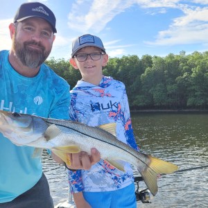 Two people holding a fish on a boat with trees in the background on a sunny day.
