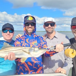 Four people smiling, holding a large fish together, with water and sky in the background.