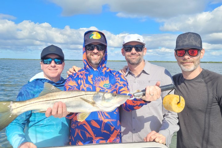 Four people smiling, holding a large fish together, with water and sky in the background.