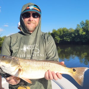 Man on a boat holding a fish, with trees and water in the background.