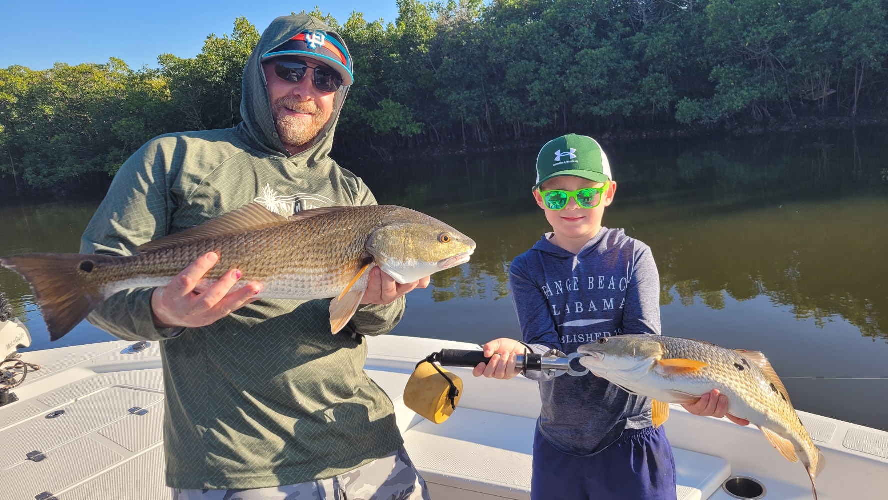 Two people on a boat holding fish in front of a tree-lined waterway.