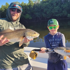 Two people on a boat holding fish in front of a tree-lined waterway.
