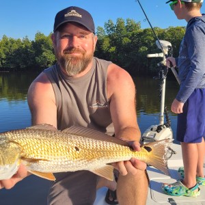 Man holding a fish on a boat, with a boy fishing in the background.