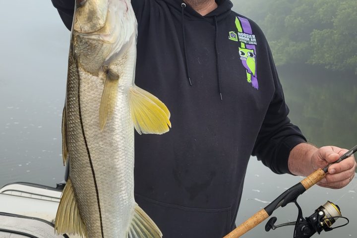 Man holding a large fish on a boat, wearing a hat and glasses.