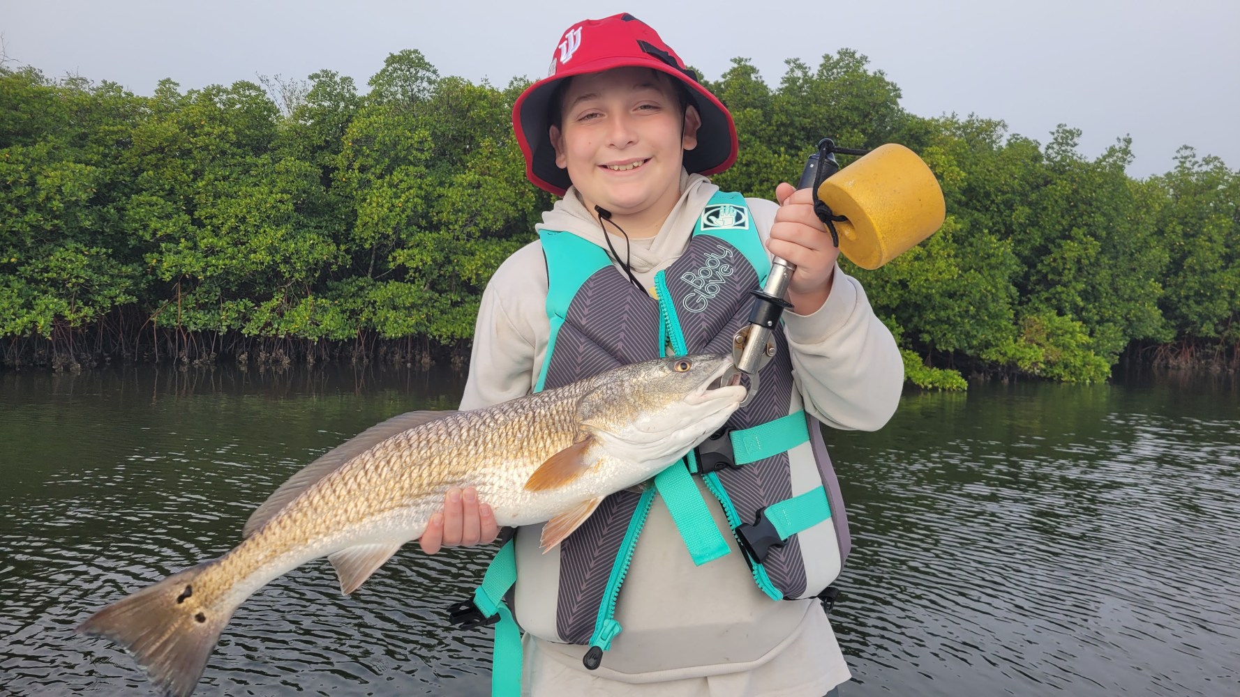 Child with red hat and life vest holding a large fish by a riverbank with trees.