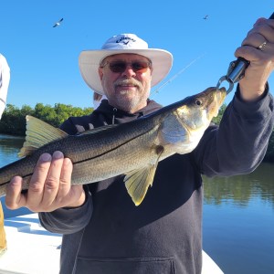 Man in hat holding a large fish on a boat with another person and trees in the background.