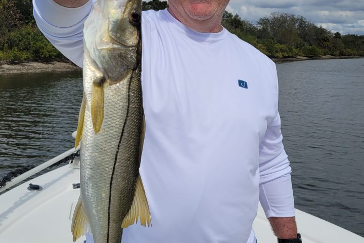 Man on a boat holding a fish up proudly, wearing sunglasses and a cap.