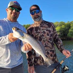 Two men on a boat holding a fish, with trees in the background.