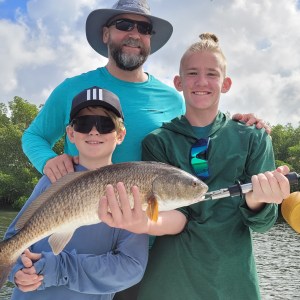 Smiling man and two boys holding a fish by the water with trees.