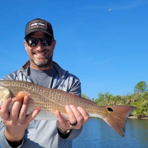Man smiling, holding a large fish by a river under a clear blue sky.