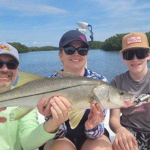Three people on a boat holding a large fish, smiling against a lake and forest backdrop.