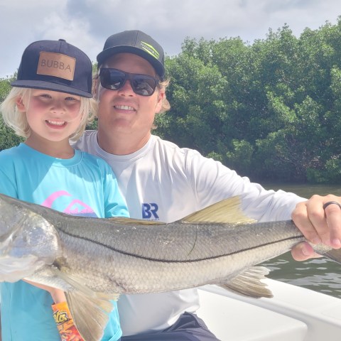 Child and adult on a boat holding a large fish, with trees in the background.