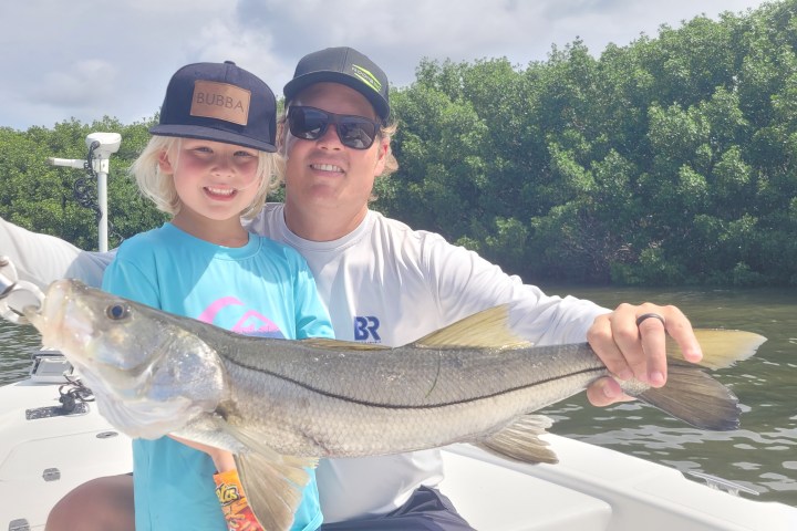Child and adult on a boat holding a large fish, with trees in the background.