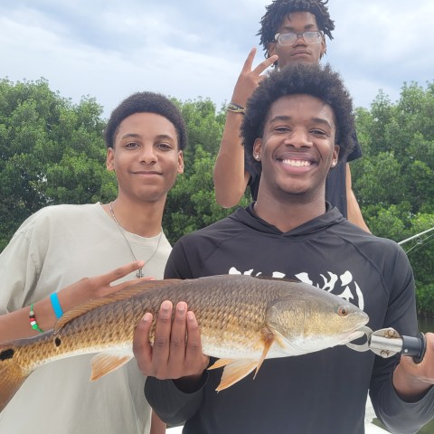 Three people on a boat, one holding a large fish, green foliage in the background.