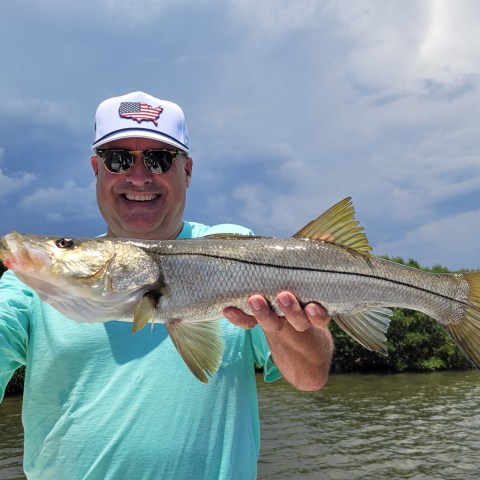 Man smiling, holding a fish by a river, wearing sunglasses and a cap.