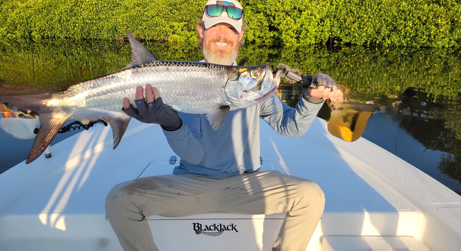 Person holding a Tarpon sitting on a boat, greenery in background.