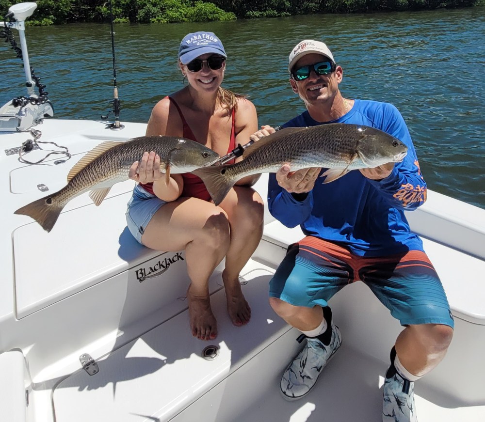 Two people on a boat holding large fish, with water and greenery in the background.